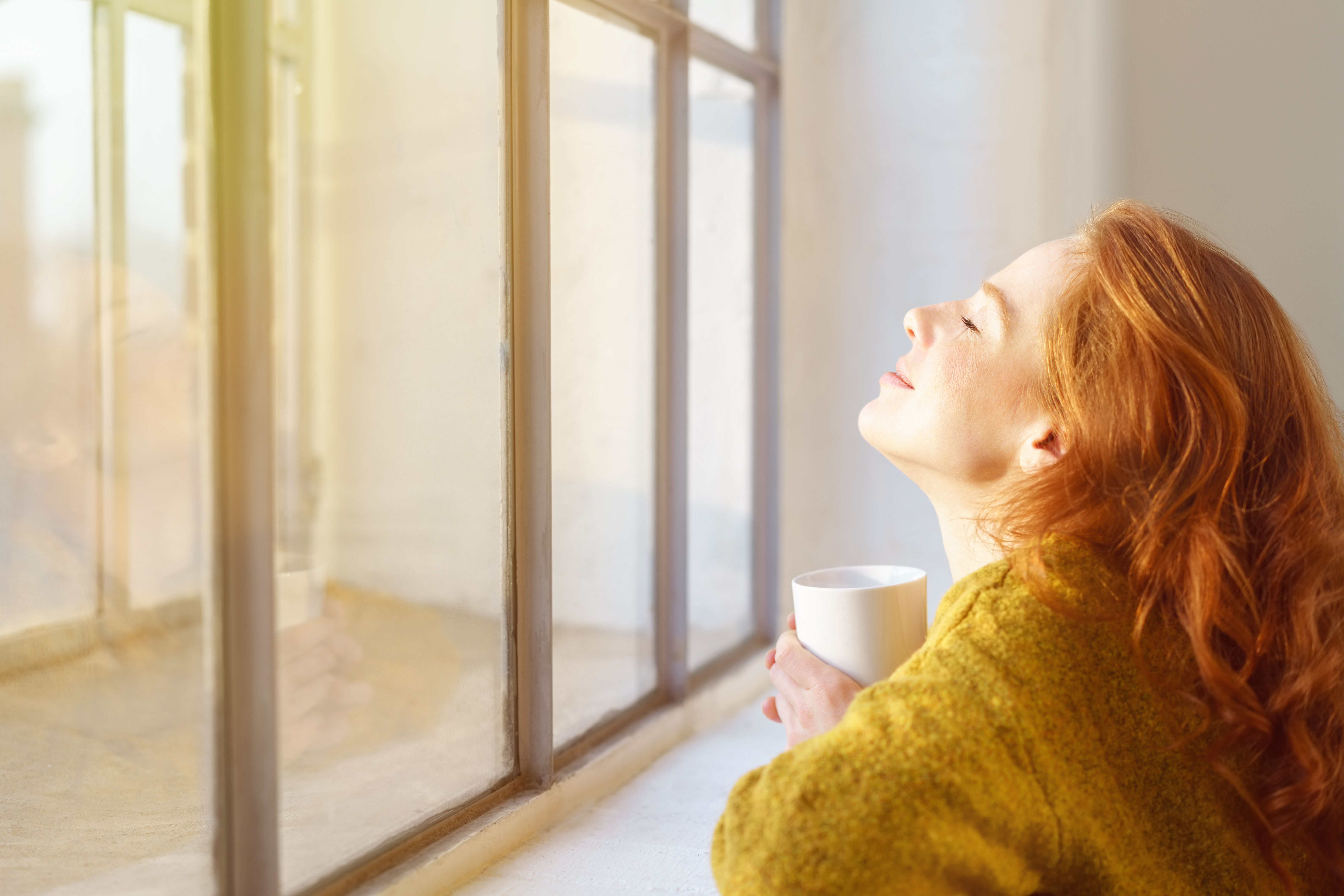 Entspannte Frau, die mit Tasse vor einem Fenster sitzt und die Sonne genießt